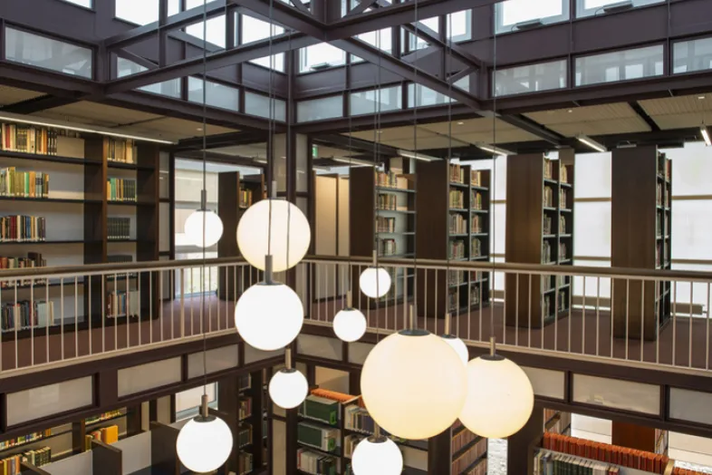 Au premier plan, les luminaires ronds de la bibliothèque, avec au 2ème plan, la balustrade et la structure métallique du plafond, les fenêtres et les rayonnages avec les livres.